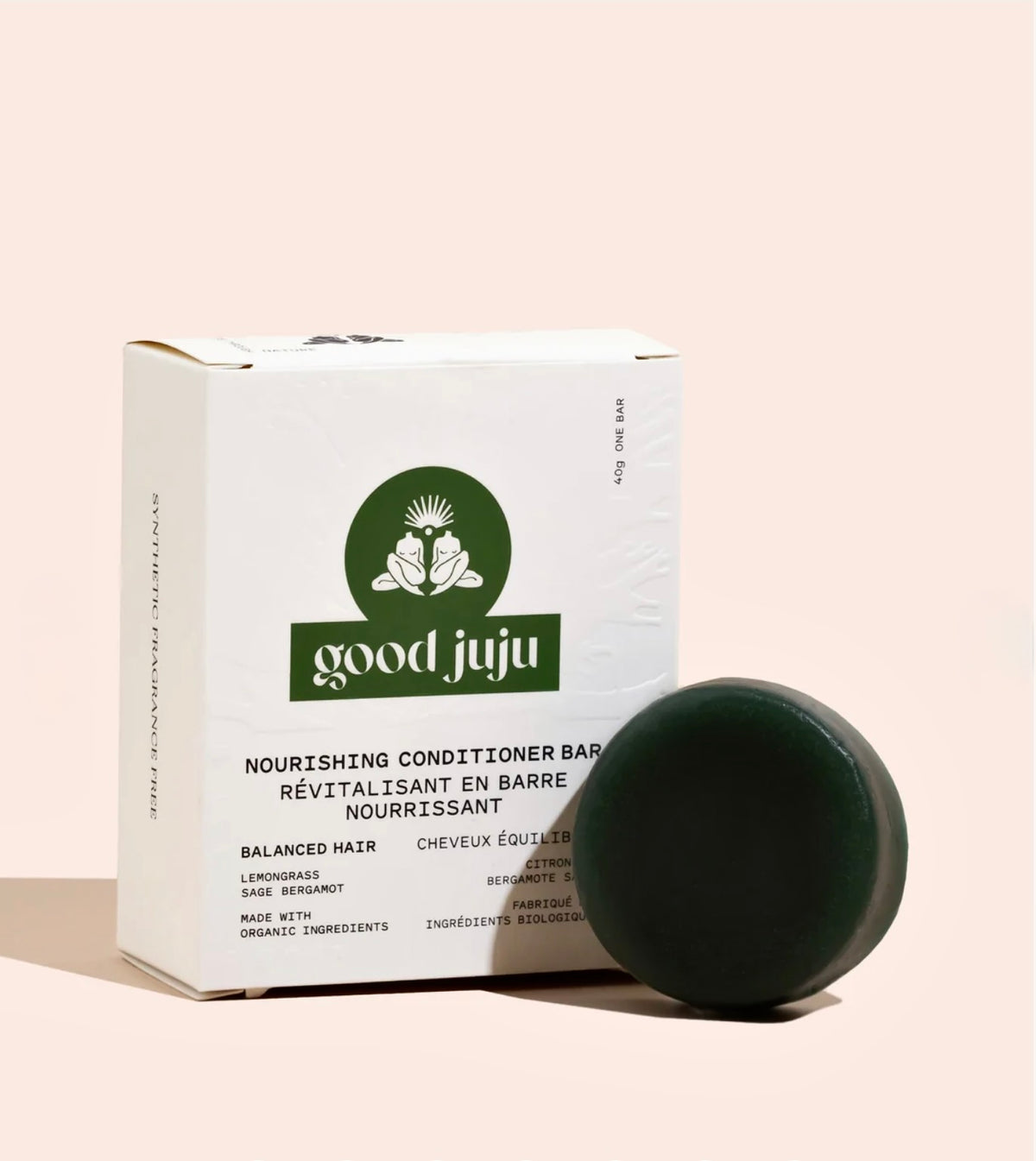 A dark green oval-shaped hair conditioner bar in front of its white cardboard packaging box, which features the green 'good juju' brand logo, set against a solid pale pink background.