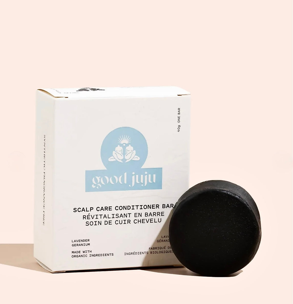 A round black scalp care conditioner bar next to its white and light blue Good Juju box on a solid pale pink background.