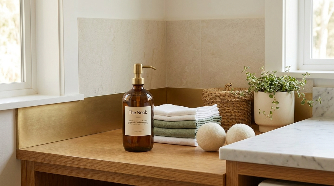 Refillable amber glass bottle of natural non-toxic laundry detergent on a honey-oak laundry bench with folded sage-green cloths and wool dryer balls