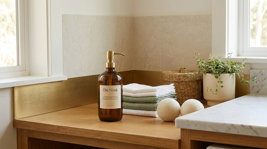 Refillable amber glass bottle of natural non-toxic laundry detergent on a honey-oak laundry bench with folded sage-green cloths and wool dryer balls