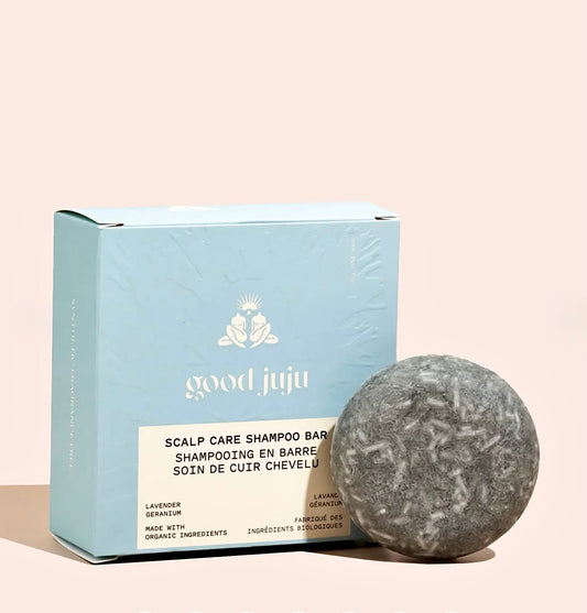 A light blue box of Good Juju Scalp Care Shampoo Bar next to a single round gray shampoo bar on a pale pink background.